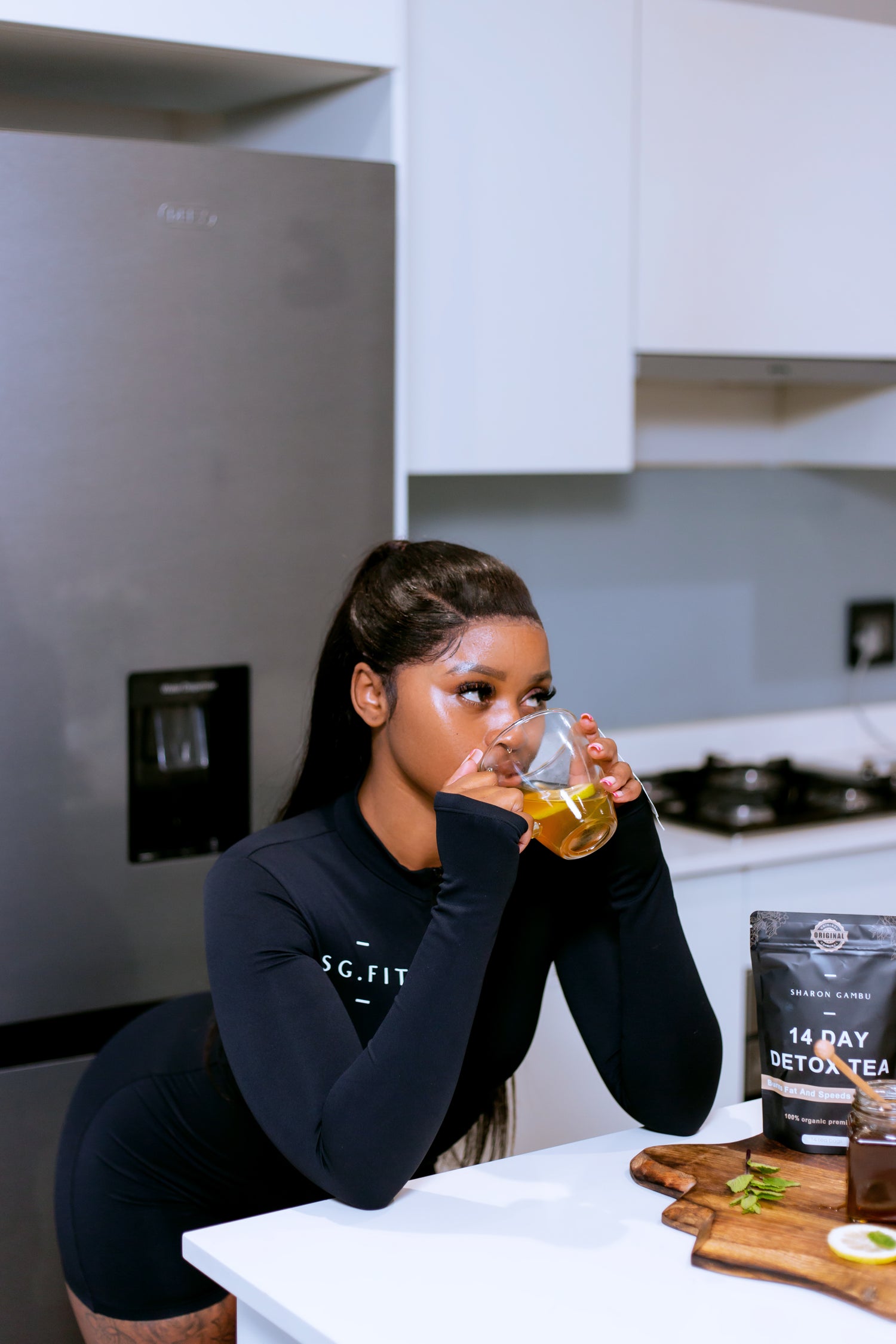 A health-conscious woman in a sleek SG.FIT black athletic top enjoys a calming sip of golden-hued detox tea in a modern kitchen setting. The countertop is clean and white, reflecting a lifestyle of clarity and purity. A wooden board, fresh mint, a sliced lemon, and a jar of honey accompany the package of Sharon Gambu 14 Day Detox Tea, highlighting the natural and organic elements of the tea experience.